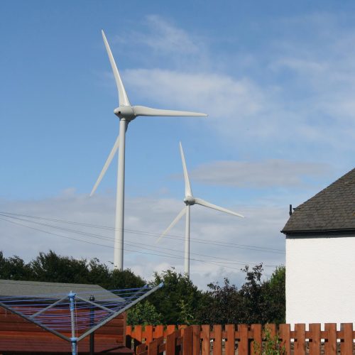 Two wind turbines behind a residential area with a house corner and a clothesline visible under a clear blue sky.