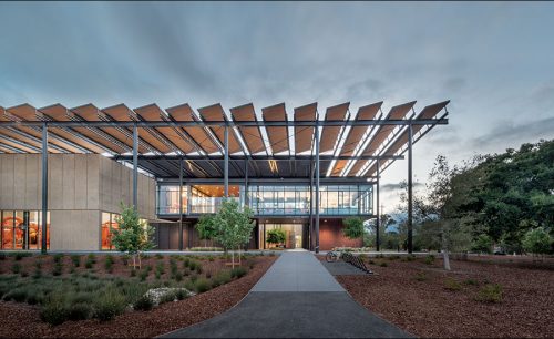 Modern educational building with a large glass facade and a prominent geometric roof, surrounded by landscaped gardens at dusk.