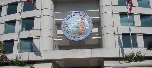 Exterior of a modern building with the great seal of california displayed prominently above the entrance, flanked by u.s. and california flags.