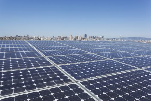 Vast solar panel array in the foreground with a city skyline and clear blue sky in the background.