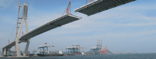 A partially constructed suspension bridge over a body of water with cranes and shipping containers in the background.