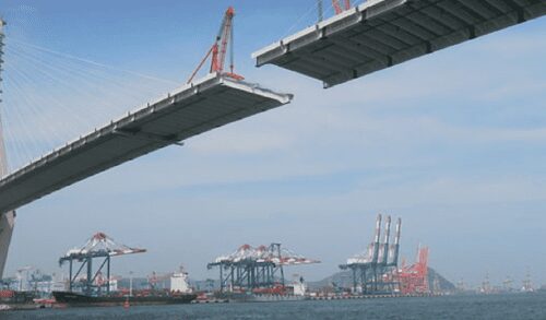 A partially constructed suspension bridge over a body of water with cranes and shipping containers in the background.