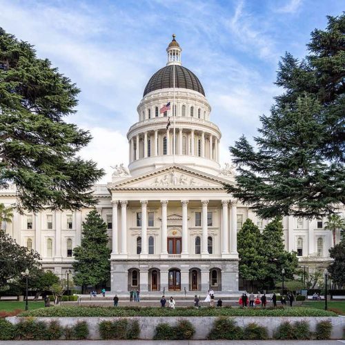 Exterior view of the california state capitol building with a domed roof, surrounded by trees and people walking in the foreground.