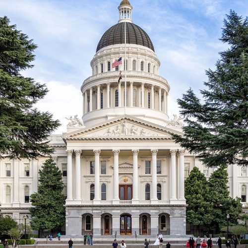 Front view of the california state capitol building, featuring its distinctive white facade and dome, flanked by trees, with people walking in front.