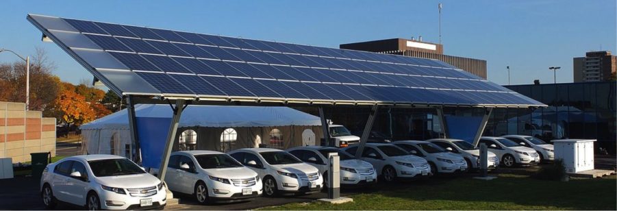 Large solar panels mounted over a carport shading several parked cars, with buildings in the background under a clear blue sky.