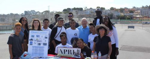 A diverse group of people posing behind an informational booth with posters at a community clean-up event.