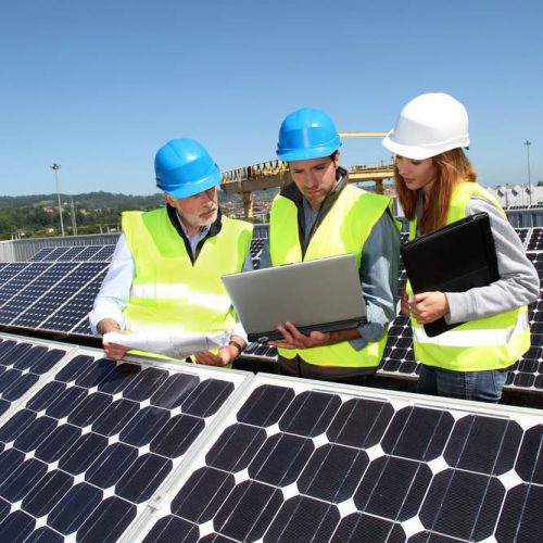 Three engineers in hard hats and high-visibility vests examining a laptop while surrounded by solar panels under a clear blue sky.