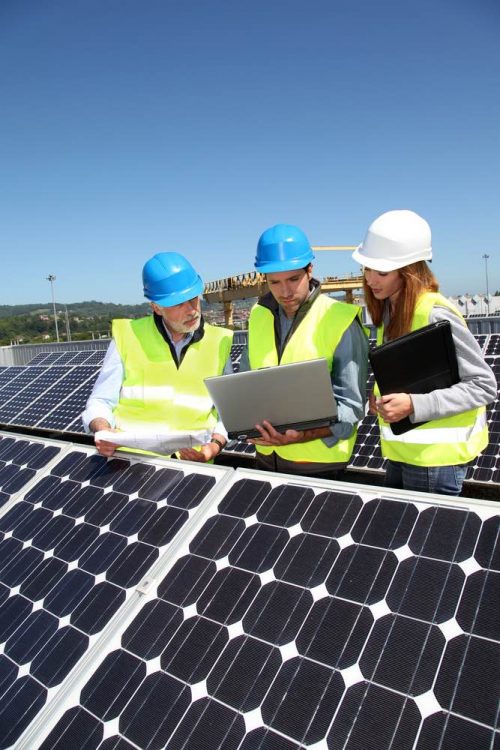 Three engineers in hard hats and high-visibility vests examining a laptop while surrounded by solar panels under a clear blue sky.