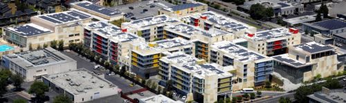 Aerial view of a modern residential complex with solar panels on rooftops, surrounded by greenery and roads.