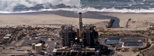 Aerial view of a coastal industrial plant with a tall striped smokestack near breaking ocean waves.