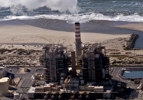 Aerial view of a coastal industrial plant with a tall striped smokestack near breaking ocean waves.
