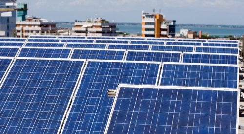 Rows of blue solar panels on a rooftop against a backdrop of urban buildings and a distant body of water.