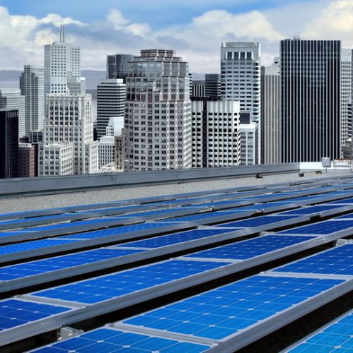 Solar panels in the foreground with a modern city skyline under a cloudy sky in the background.