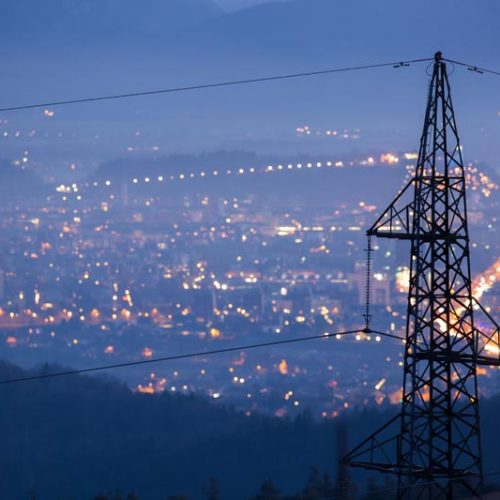 Electricity pylon silhouetted against a night cityscape with twinkling lights and a hazy atmosphere.