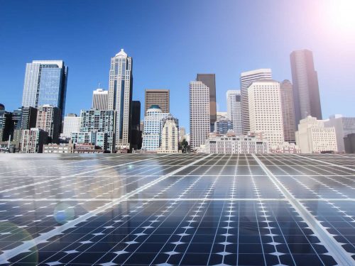 Solar panels in the foreground with a city skyline under a clear blue sky and bright sun.