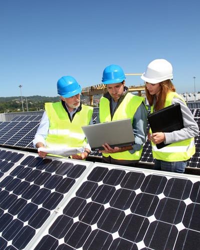 Three engineers in hard hats using a laptop and taking notes on clipboards at a large solar panel array on a sunny day.