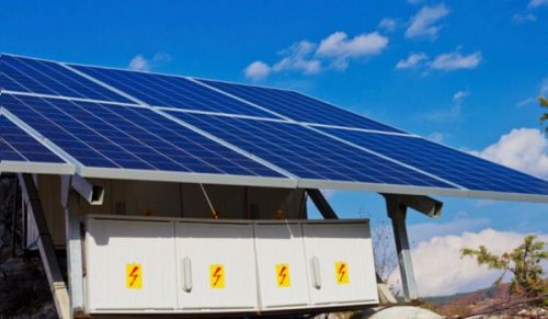 Large solar panels mounted on top of storage batteries with clear blue sky and mountains in the background.