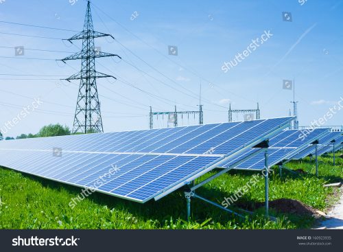 Solar panels array in a field with an electricity pylon and substation in the background under a clear blue sky.