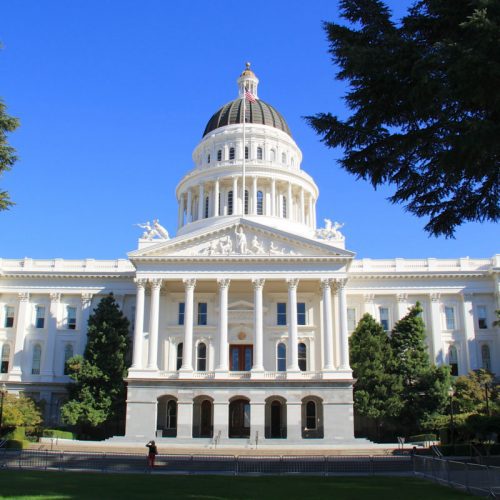 The california state capitol building in sacramento, viewed from the front on a sunny day, framed by lush green trees.