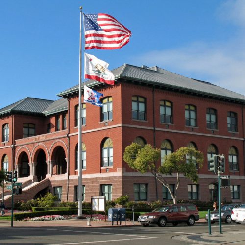Red brick public building with a large staircase and three flags, including the american flag, flying on a flagpole in front.