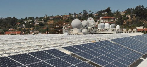 Solar panels in the foreground with satellite dishes and residential houses on a hill in the background under a clear blue sky.