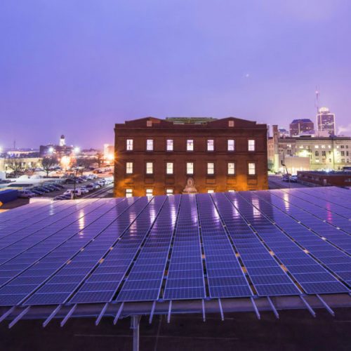 A cityscape at twilight featuring a large solar panel array in the foreground with an old brick building and city lights in the background.