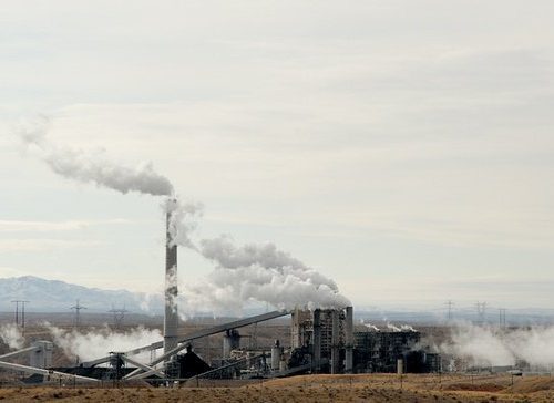Industrial plant in a desert landscape emitting smoke from stacks against a clear sky, with mountains in the background.