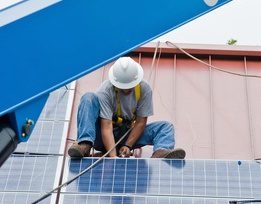 A worker in a hard hat and safety harness installs solar panels on a rooftop.