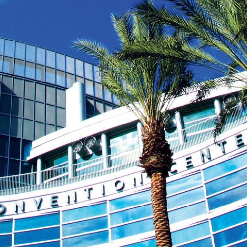 Modern glass facade of the anaheim convention center with tall palm trees in the foreground under a clear blue sky.