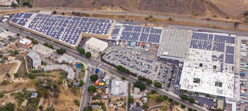 Aerial view of an industrial area with large buildings covered in solar panels, adjacent to a parking lot and roadway.
