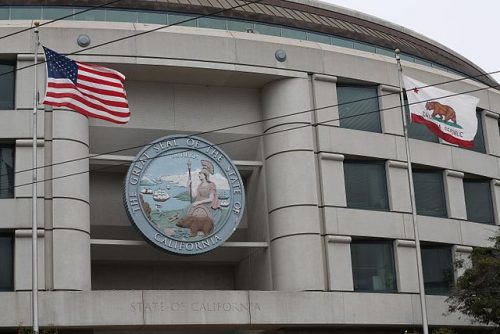 Exterior view of a civic building in california with an american flag and the california state flag flying above. the building features a large emblem of the great seal of california.