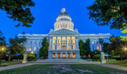 Nighttime view of a lit-up state capitol building with a large dome, surrounded by green lawns and trees under a twilight sky.