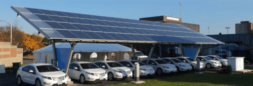 A solar panel carport covering a row of parked cars, with buildings and trees in the background on a clear day.