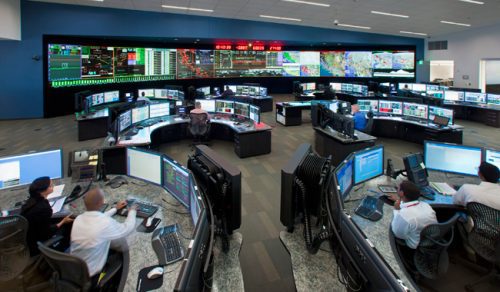 High-tech control room with multiple workers at computer stations monitoring large digital screens displaying maps and data.