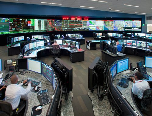 High-tech control room with multiple workers at computer stations monitoring large digital screens displaying maps and data.