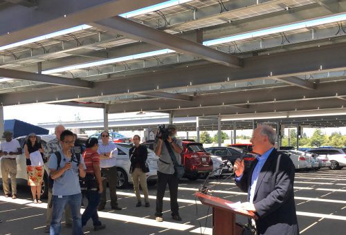 A man speaks at a podium in a solar-paneled parking lot, addressing an audience including a cameraman and several listeners.