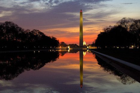 Washington monument at sunset, reflecting in the still water of the reflecting pool with glowing lights and colorful sky.