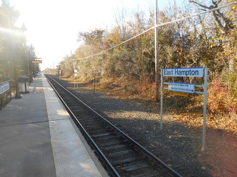 Deserted east hampton train station platform with track on a sunny day, featuring a station sign and overhead power lines.
