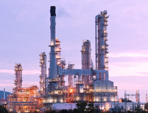 An oil refinery illuminated at twilight with tall distillation towers and complex piping, set against a soft purple sky.