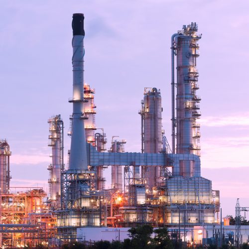 An oil refinery illuminated at twilight with tall distillation towers and complex piping, set against a soft purple sky.