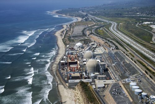 Aerial view of a coastal nuclear power plant next to a highway and beach, with expansive ocean and shoreline visible.