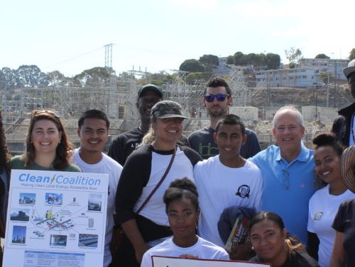 Diverse group of people posing for a photo at a waterfront, with some holding a project poster titled 