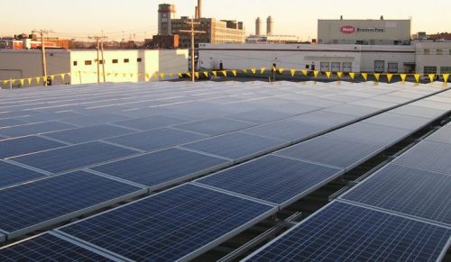 Rows of solar panels on a rooftop at sunset, with industrial buildings in the background.