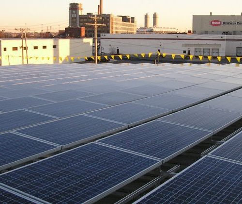 Rows of solar panels on a rooftop at sunset, with industrial buildings in the background.