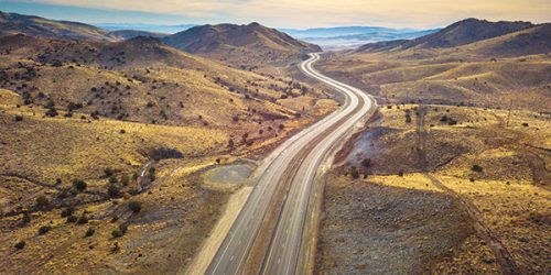 Aerial view of a winding road cutting through a hilly, arid landscape under a clear sky.