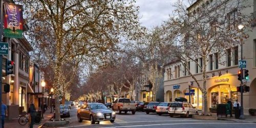 A bustling city street at dusk showing cars, pedestrians, and illuminated trees along a shopping district with vintage architecture.