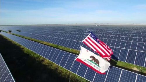 Aerial view of a vast solar farm with the u.s. and california flags displayed prominently in the foreground.