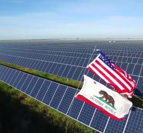 Aerial view of a vast solar farm with the u.s. and california flags displayed prominently in the foreground.