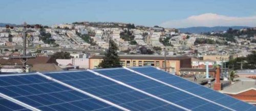 Solar panels in the foreground with a panoramic view of a hilly residential area under a clear blue sky.