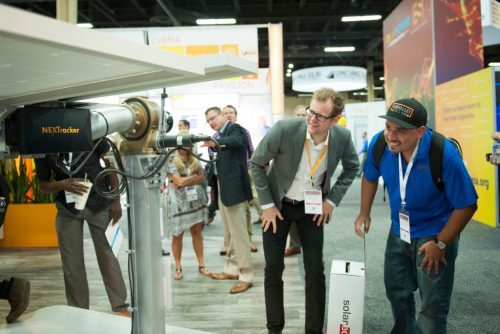 Two men examining a large camera equipment at a trade show with other attendees in the background.